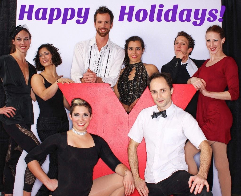 A goofy, low-budget holiday-card style image. Eight artists in formal costumes smile cheesily at the camera. At the top of the card is bright purple text that says &ldquo;Happy Holidays&rdquo;