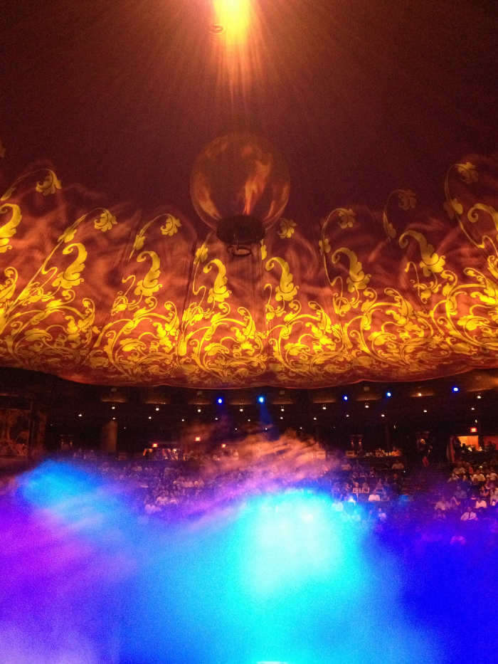 Blue fog rises up from the bottom of the stage below, which is completely surrounded by seats. Above, the top of the space is covered by bollowing red and orange fabrics, from the center of which a light shines down into the darkness